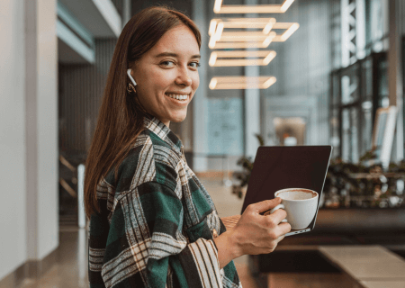 O que é assiduidade no trabalho Mulher segurando laptop em uma mão e xícara de café em outra, com fones no ouvido - Imagem de Freepik