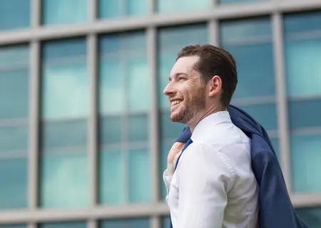 Profissional sorrindo saindo da empresa e carregando casaco nas costas com uma mão só, satisfeito pela reduação da jornada de trabalho - Imagem de Freepik