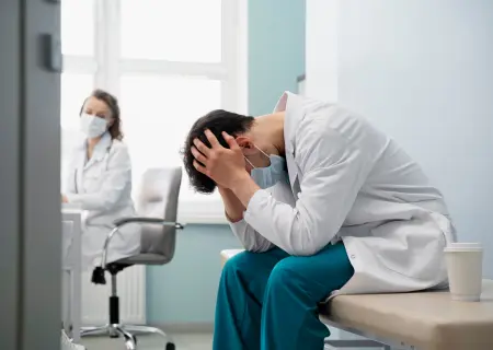 Excesso de trabalho causa Médico sentado em maca com as mãos na cabeça demonstrando cansaço por excesso de trabalho; uma colega o observa ao fundo - Imagem de Freepik